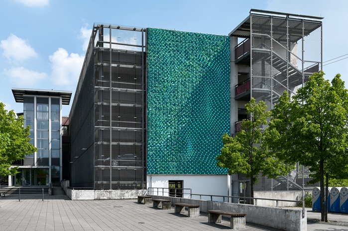 Begrünte Fassade an einem Parkhaus: eine große grüne Pflanzenwand zwischen Treppenhäusern, davor ein Platz mit Bäumen